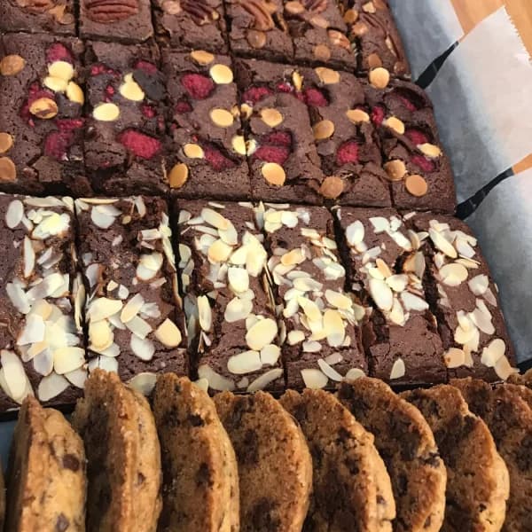 Bandeja de brownies cortados en cuadrados, unos con frambuesa, otros con chocolate blanco y otros con almendra laminada, y una fila de cookies delante