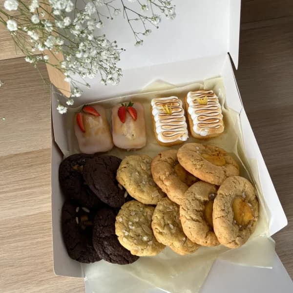 Caja blanca abierta con galletas de chocolate y de nuez, dos mini cakes glaseados y dos bocados decorados, sobre mesa clara con paniculata