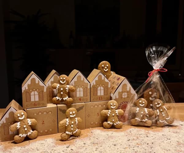 Calendario de adviento de casitas de cartón junto a galletas de jengibre con forma de muñeco decoradas con glasa blanca, una de ellas en bolsa con lazo rojo