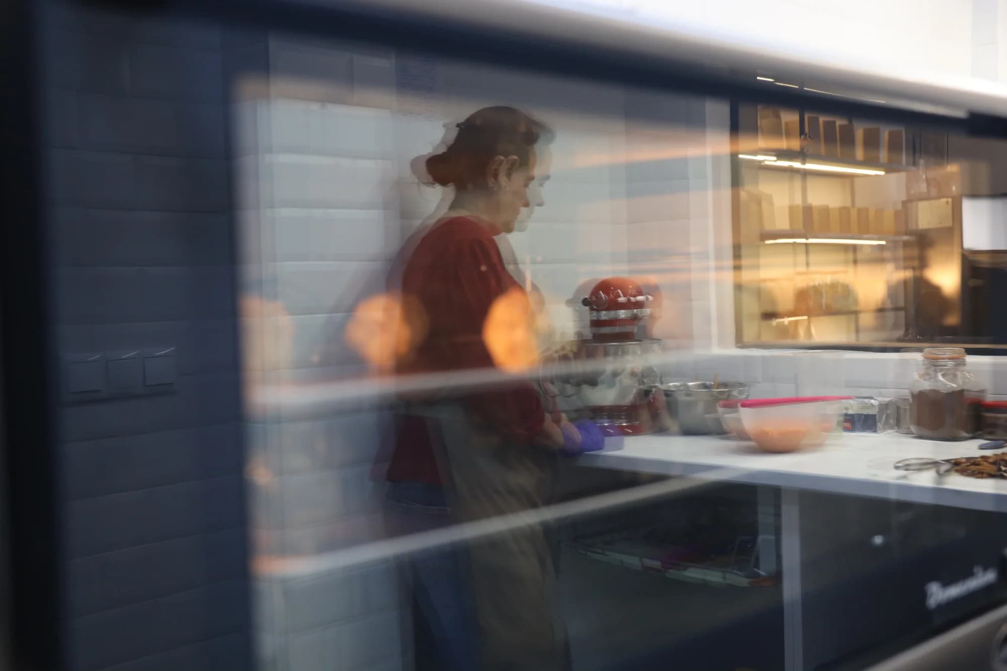 Reflejo en el cristal de la vitrina del obrador, con la repostera trabajando al fondo entre estanterías y luz cálida