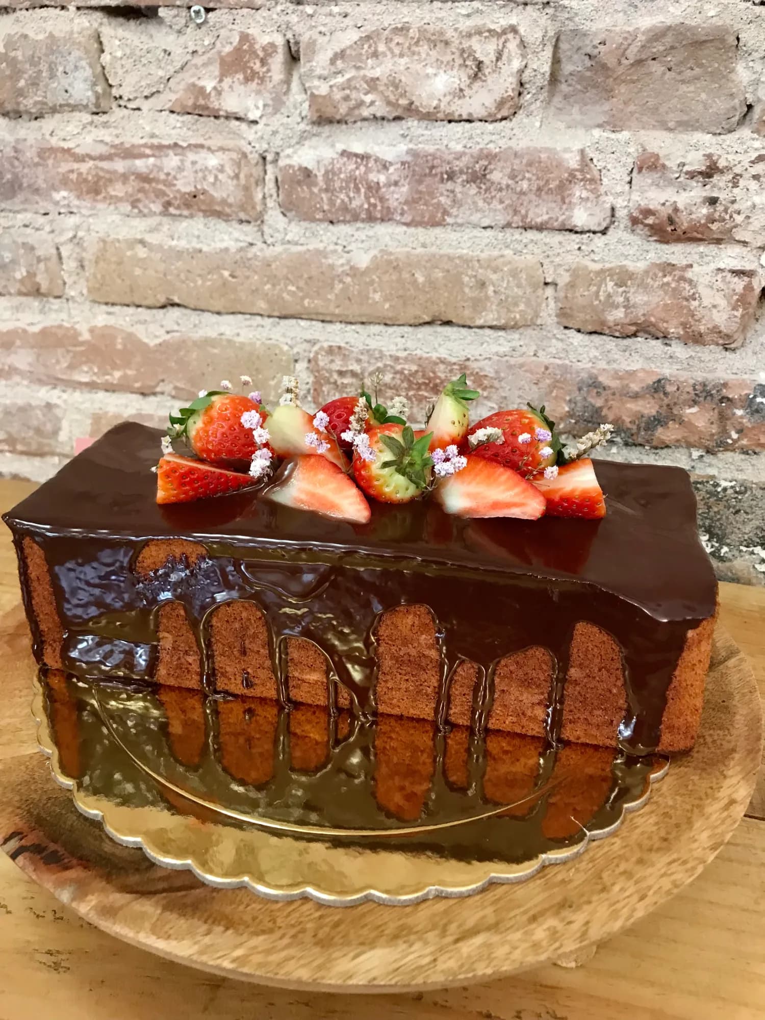 Plum cake bañado en cobertura de chocolate con fresas frescas cortadas por encima, presentado en bandeja dorada contra una pared de ladrillo visto