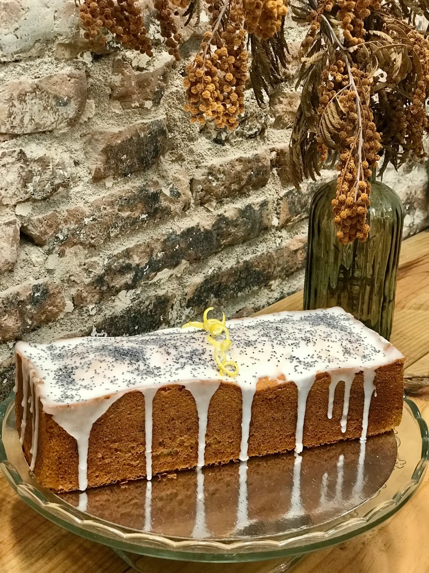 Plum cake de limón y amapola con glaseado blanco cayendo por los lados y ralladura de limón, sobre plato de cristal ante una pared de piedra