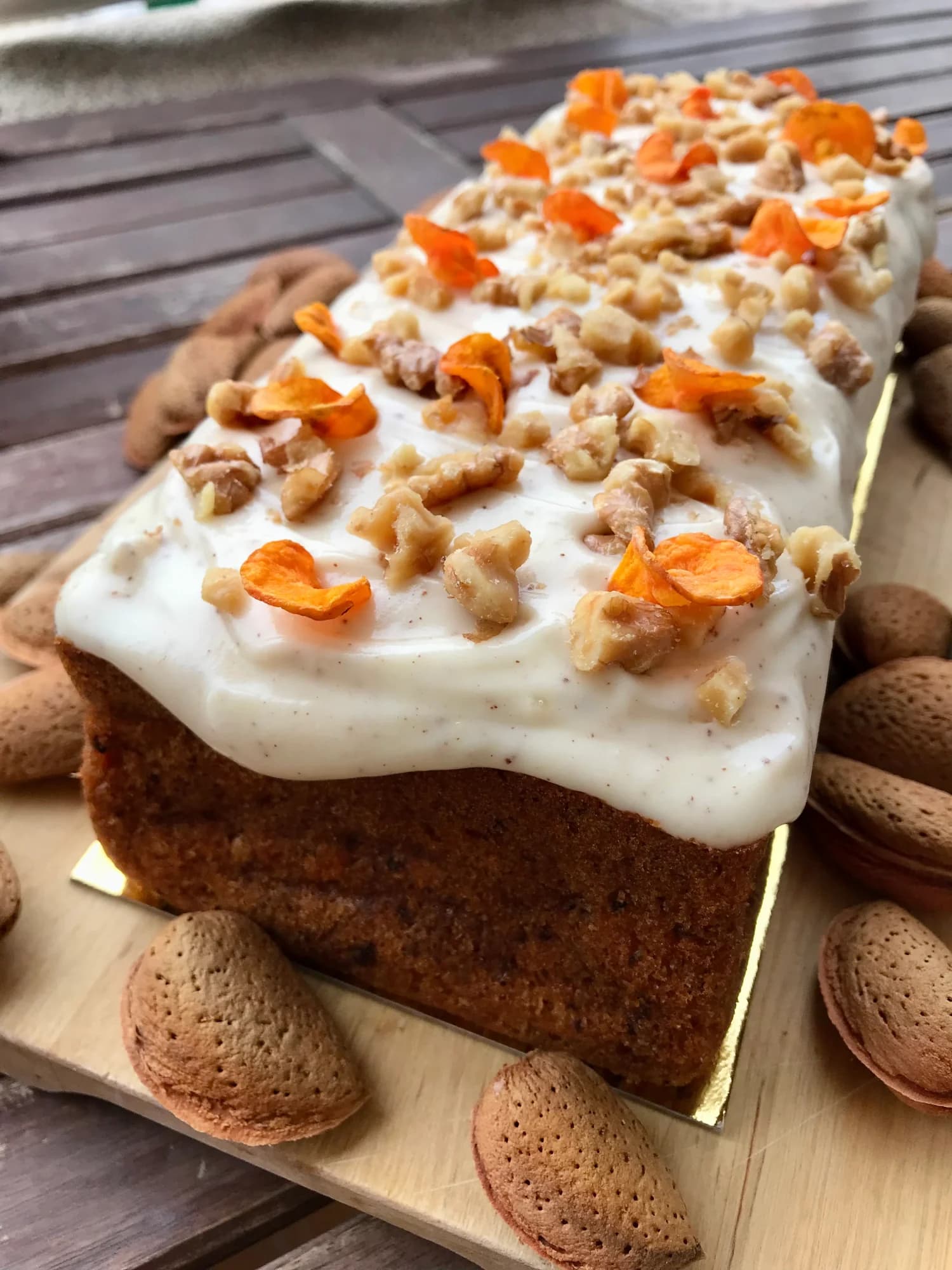 Plum cake de zanahoria cubierto de frosting de queso, nueces troceadas y pétalos de naranja deshidratada, sobre tabla de madera rodeado de almendras