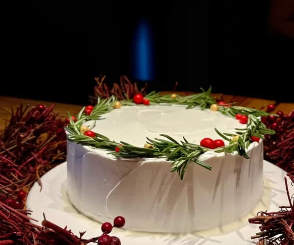 Tarta blanca con una corona de romero fresco y arándanos rojos bordeando la superficie, sobre un mantel con decoración navideña