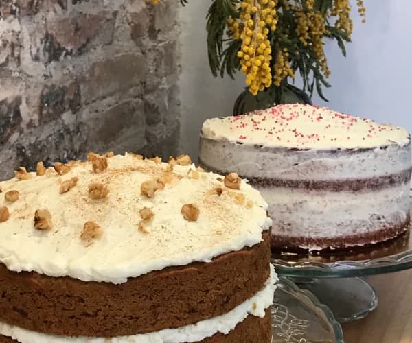 Dos tartas juntas, una de zanahoria con frosting y nueces y detrás una red velvet de bizcochos vistos, sobre pie de cristal y con mimosas al fondo