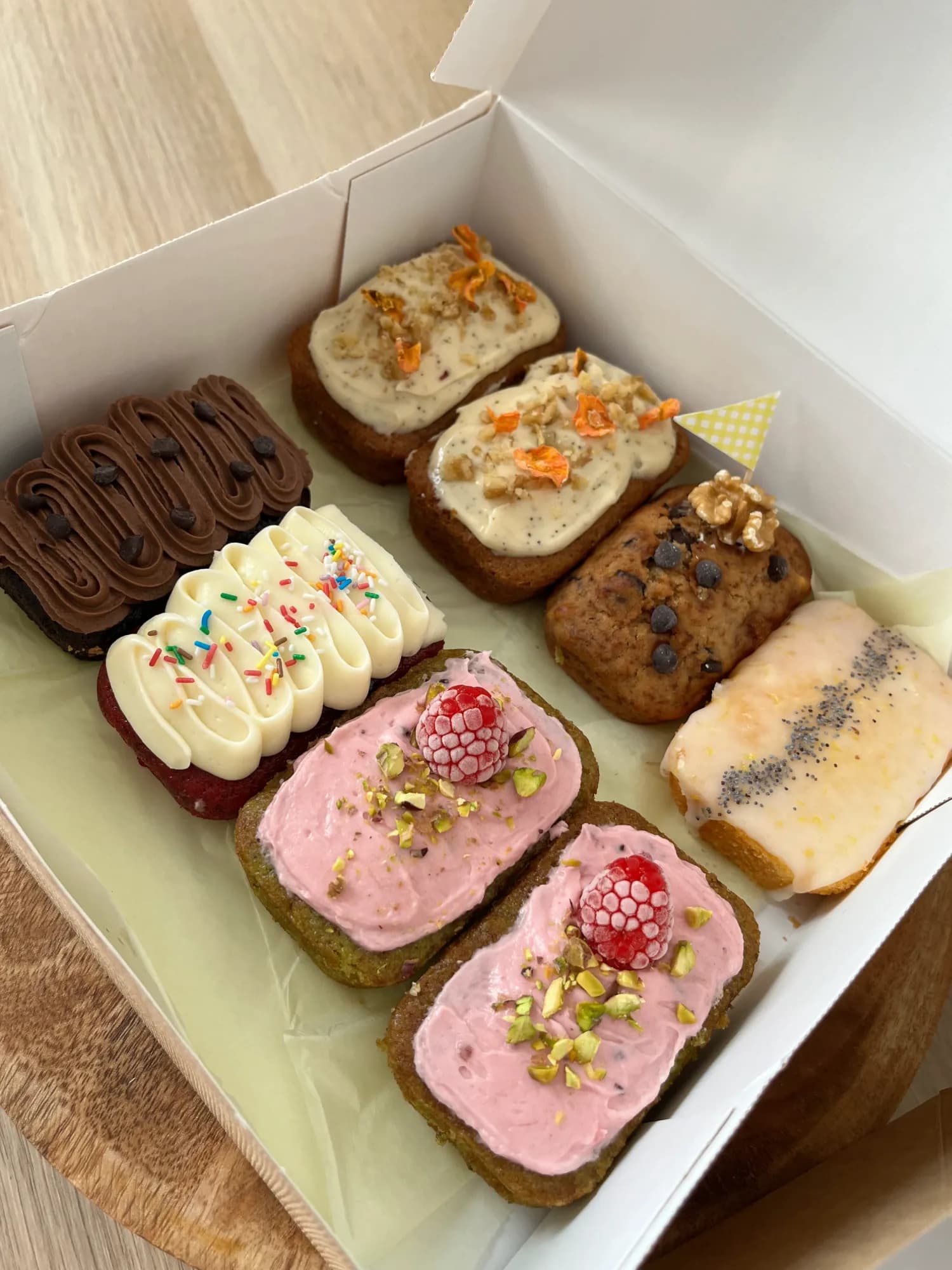 Caja de cartón abierta con ocho mini plum cakes distintos: chocolate, vainilla con fideos de colores, frambuesa y pistacho, zanahoria y limón