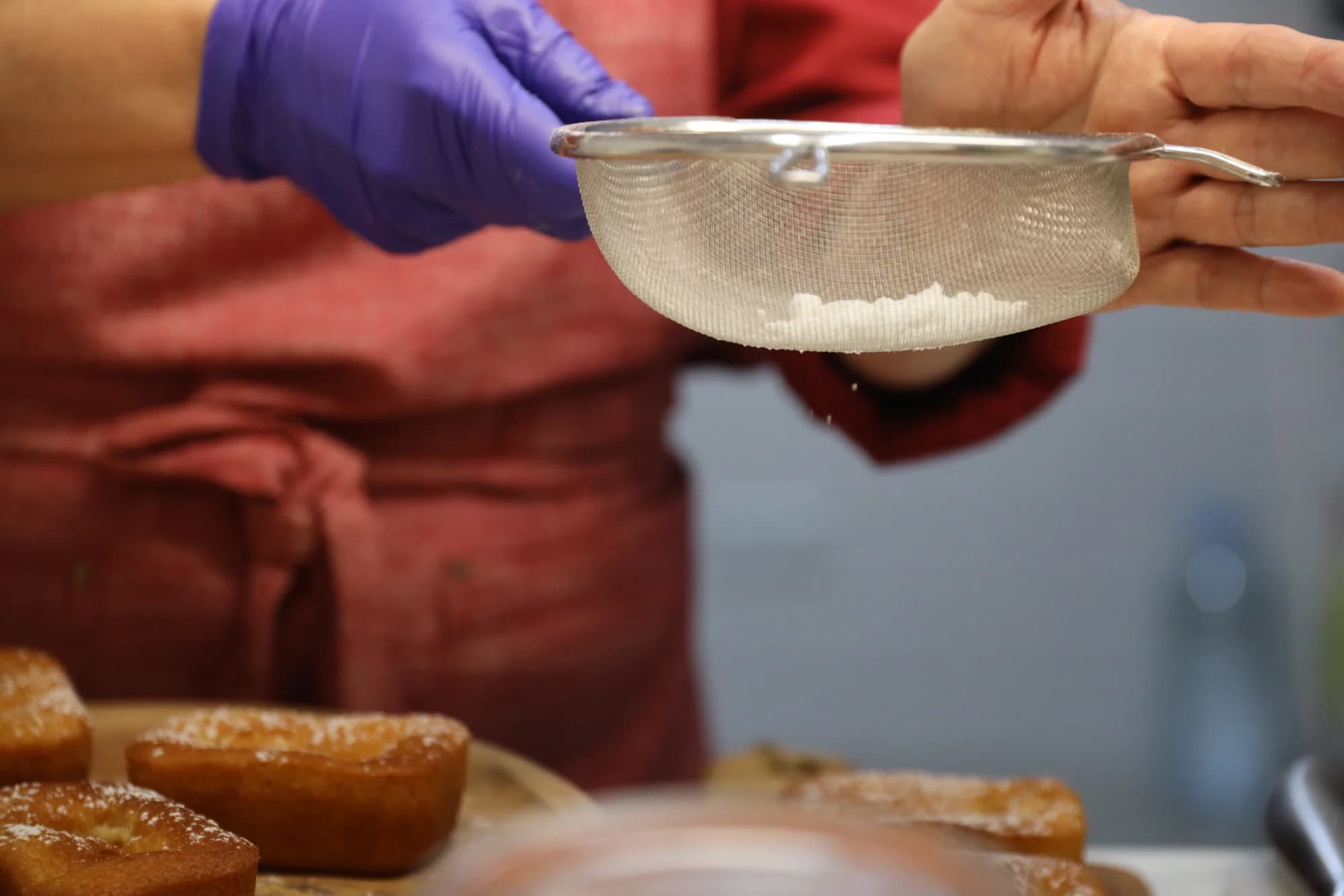 Primer plano de unas manos con guantes sosteniendo un colador con azúcar glas sobre bizcochos recién horneados