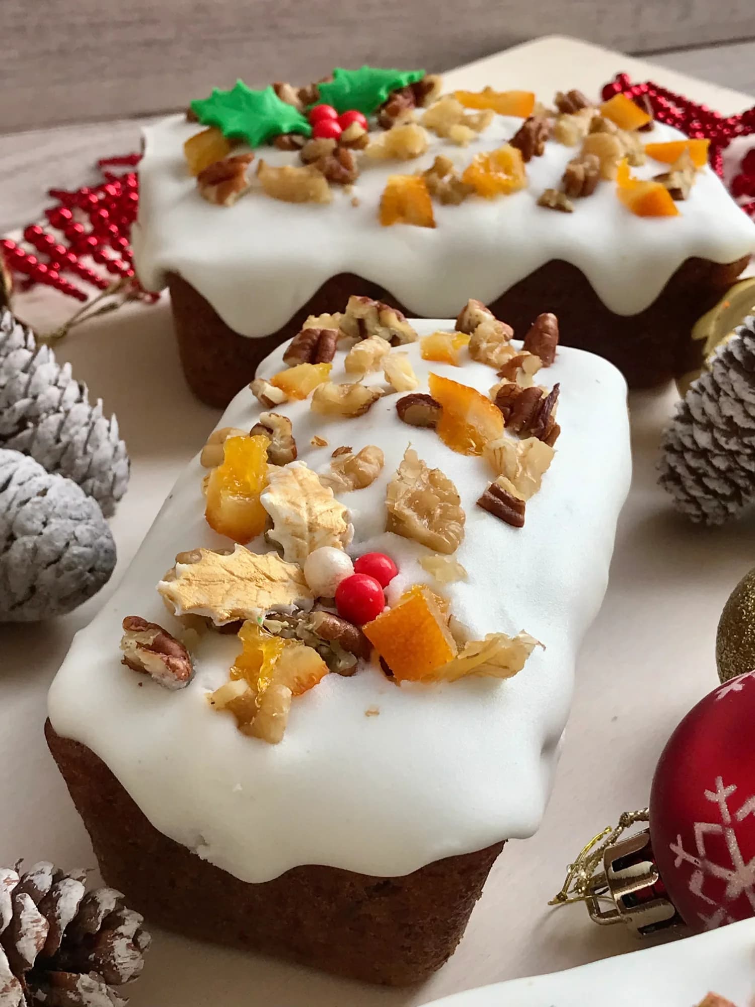 Dos plum cakes navideños glaseados en blanco, decorados con nueces, pacanas y naranja confitada, entre piñas y bolas de Navidad